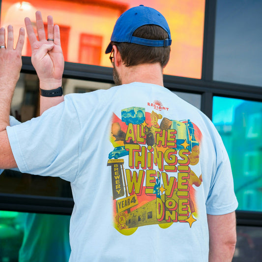 A man wearing a light blue Radiant Beer Co. t-shirt and a royal blue baseball cap faces a reflective glass wall. The back of his shirt features a vibrant, collage-style graphic with the text “ALL THE THINGS WE’VE DONE” in bold red and yellow lettering. The design includes beer cans, brewery tanks, a Radiant sign, and other illustrated elements celebrating Radiant’s “Year 4.” The Radiant Beer Co. logo appears at the top of the shirt.