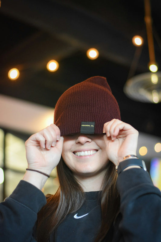 Smiling woman indoors playfully pulling a maroon beanie over her eyes, with a visible tag on the hat that reads “ANAHEIM, CA” in small yellow