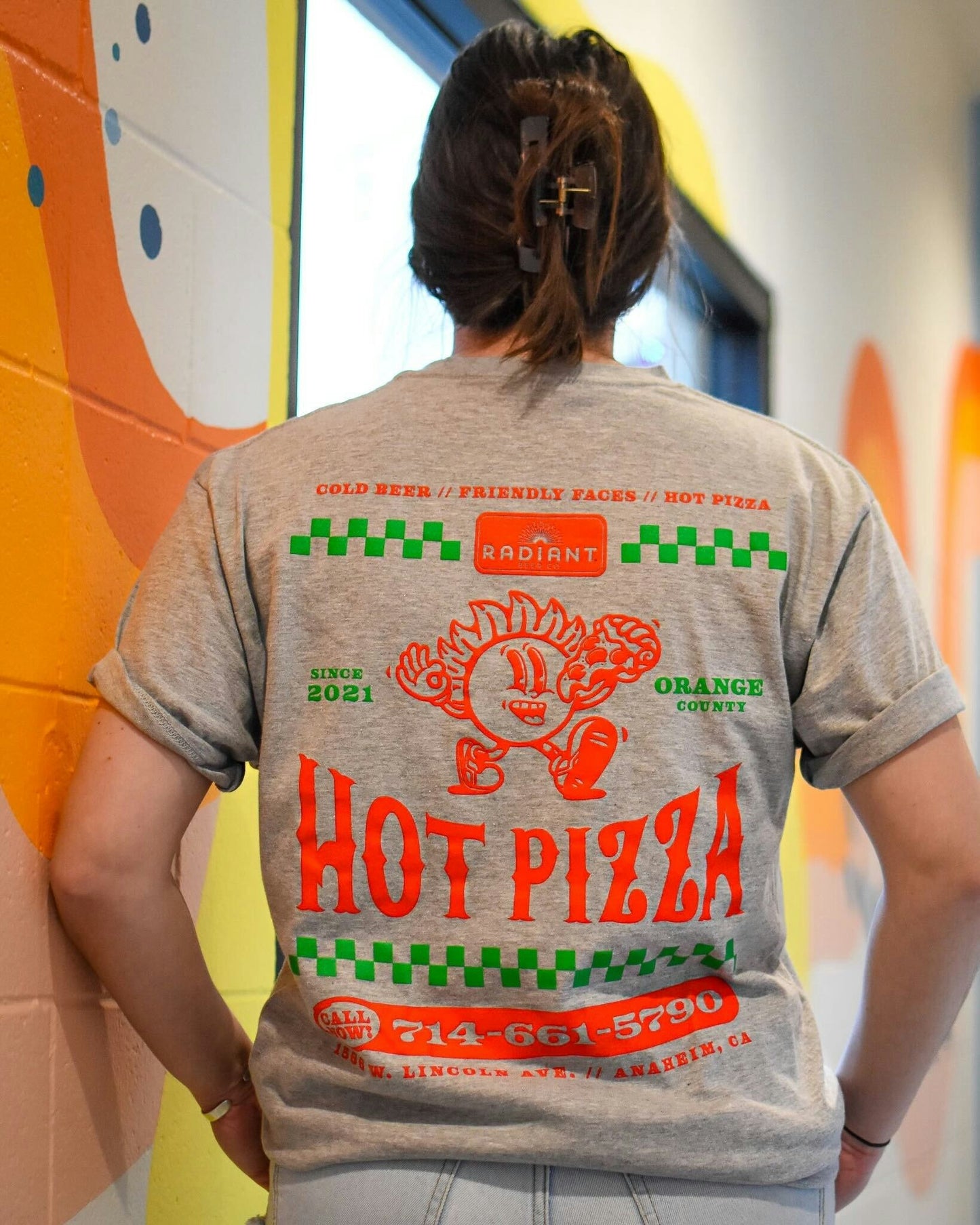 Back view of a person wearing a heather gray Radiant Beer Co. t-shirt with a colorful, retro-style design. The shirt features bold red text that says “HOT PIZZA,” a cartoon flame character, green checkered accents, and phrases like “Cold Beer // Friendly Faces // Hot Pizza,” “Since 2021,” and “Orange County.” It also displays a phone number and address for Radiant Beer Co. in Anaheim, California. The person is standing indoors against a painted wall.