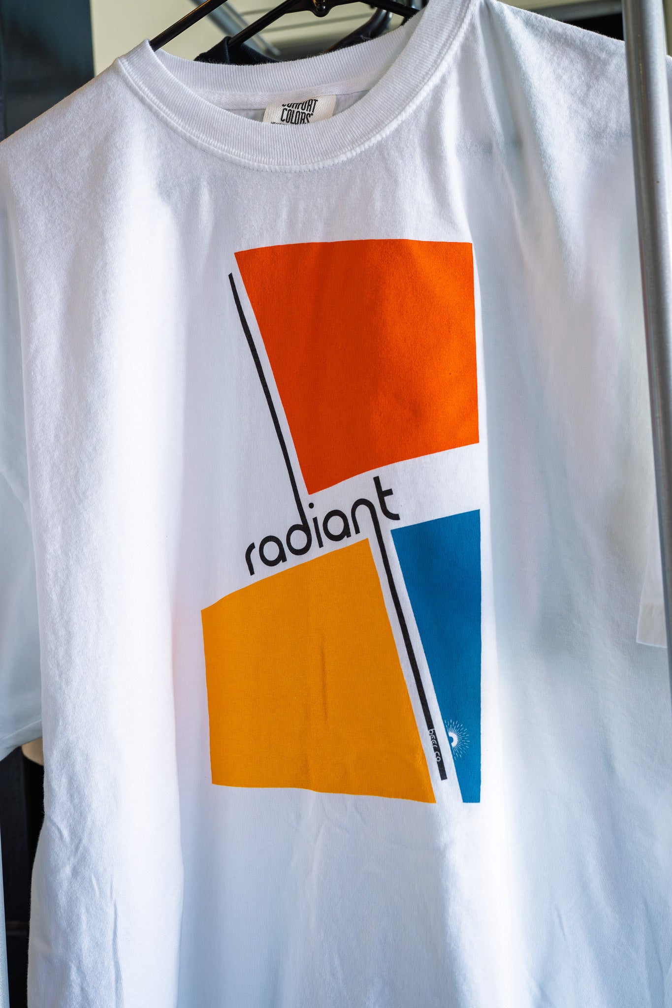 White t-shirt with colorful geometric design and 'radiant' text on a hanger.