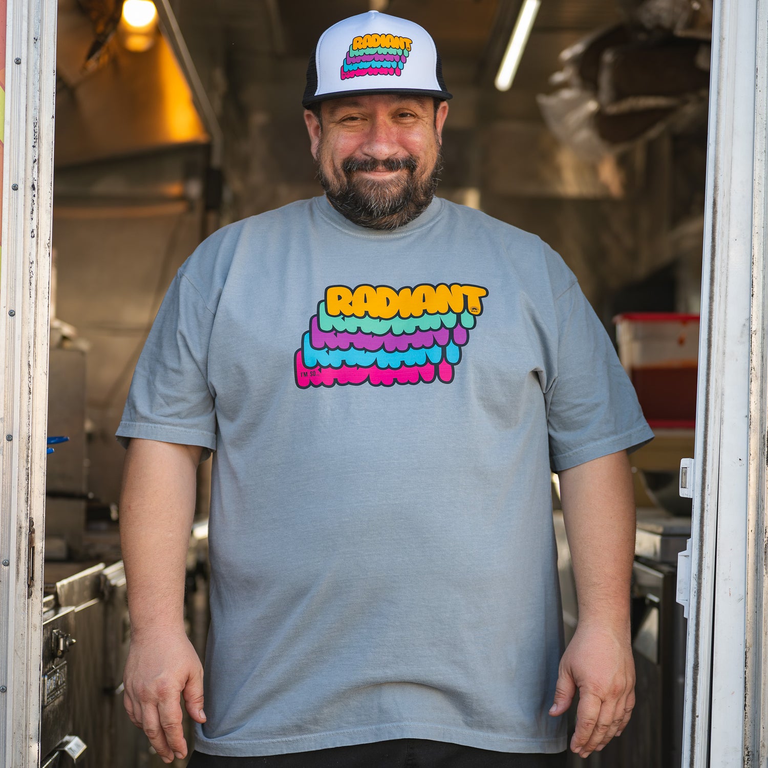 Man wearing a gray t-shirt with colorful text and a rainbow cap inside a kitchen.