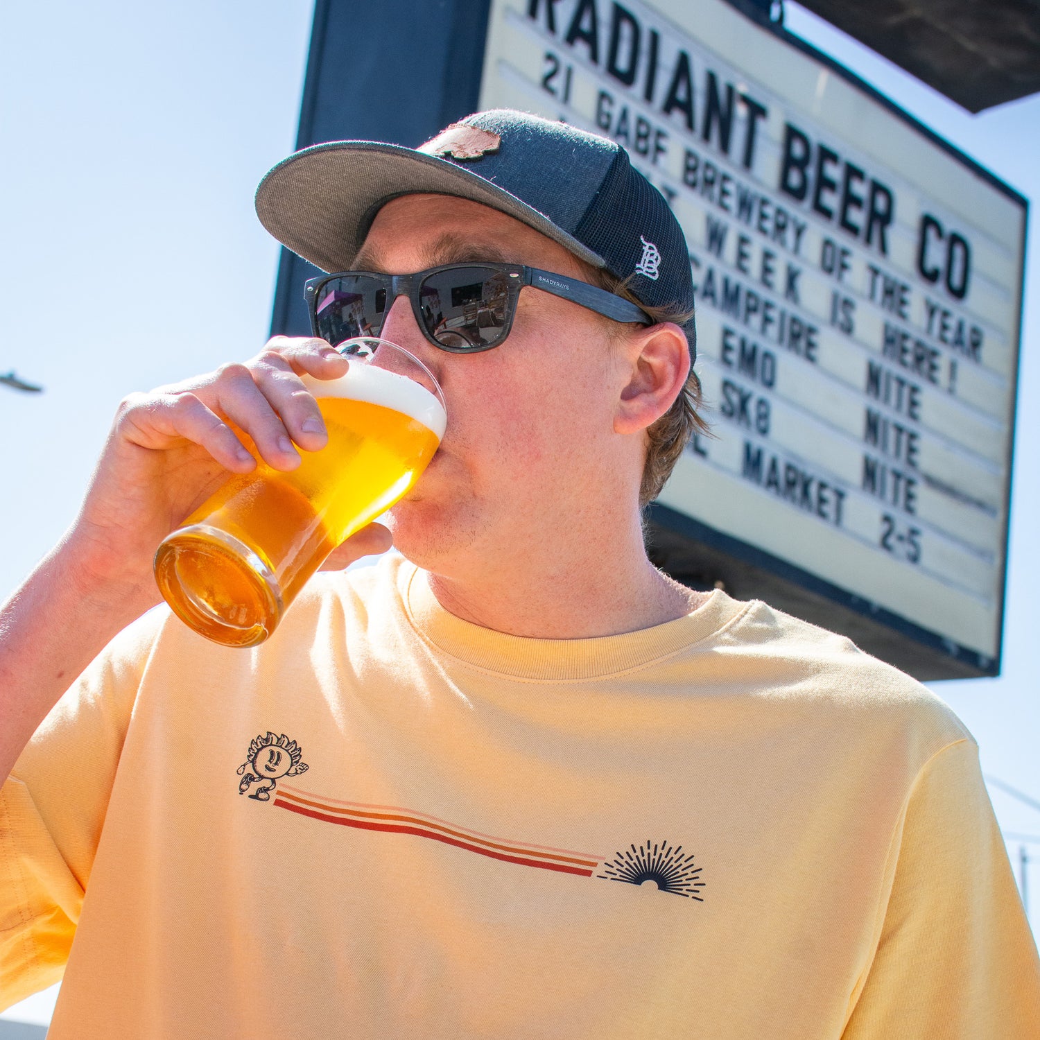 Man wearing sunglasses and a cap, holding a pint of beer while modeling a soft yellow Radiant Beer Co. t-shirt with a minimalist sunbeam logo and horizontal red-orange stripe design across the chest, photographed outdoors in bright natural light.