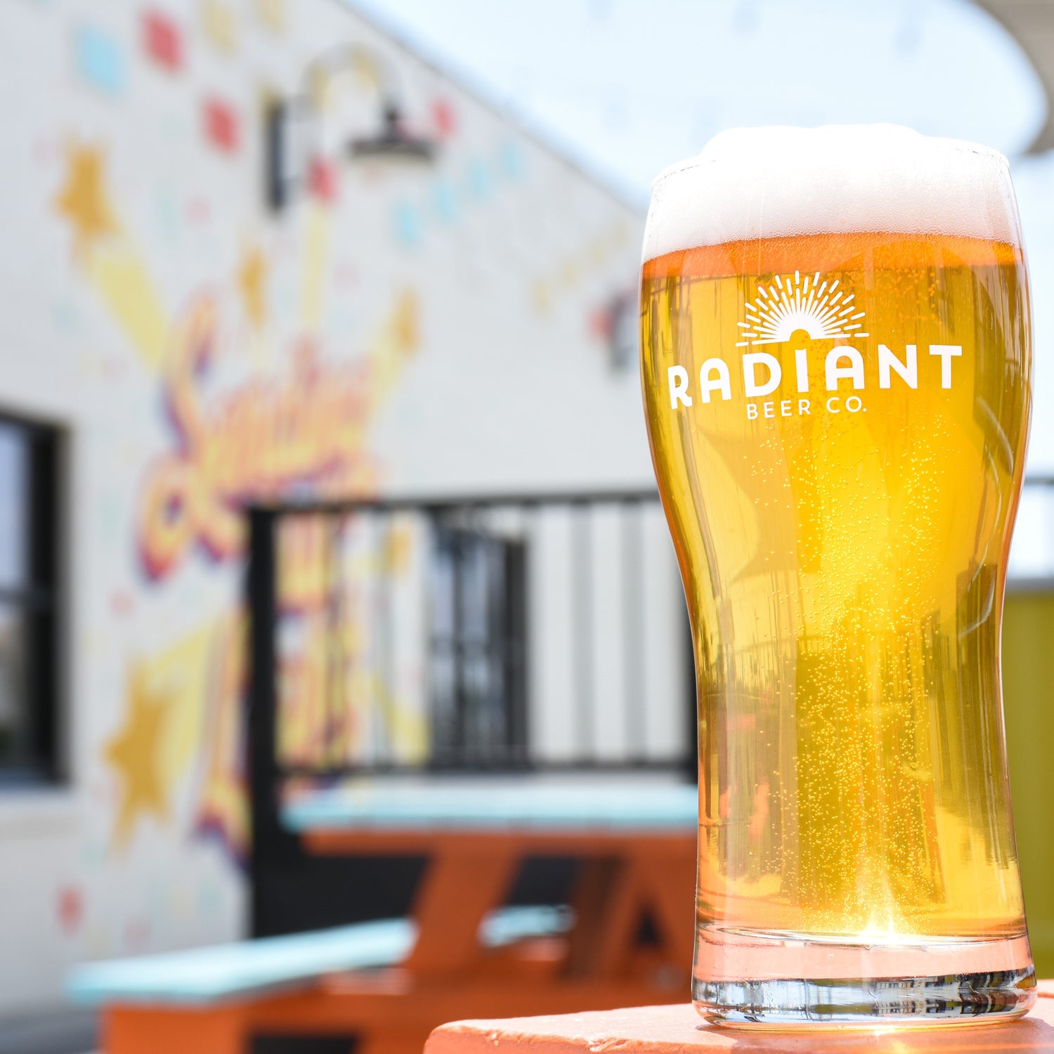 A lager with a fluffy white head sits on an orange picnic table on the patio with the mural in soft focus in the backdrop. 