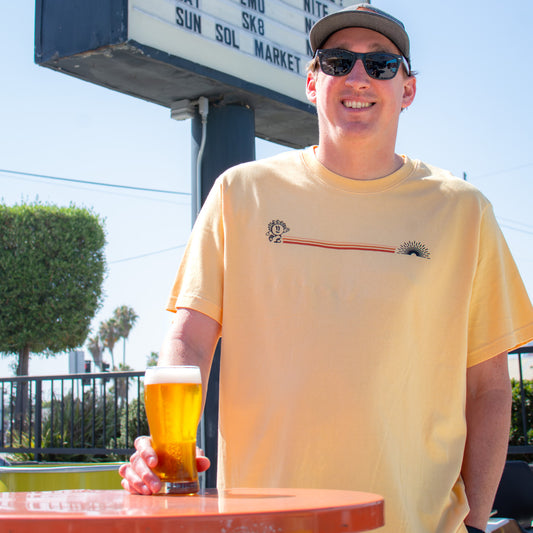 Man wearing sunglasses and a cap, holding a pint of beer while modeling a soft yellow Radiant Beer Co. t-shirt with a minimalist sunbeam logo and horizontal red-orange stripe design across the chest, photographed outdoors in bright natural light.